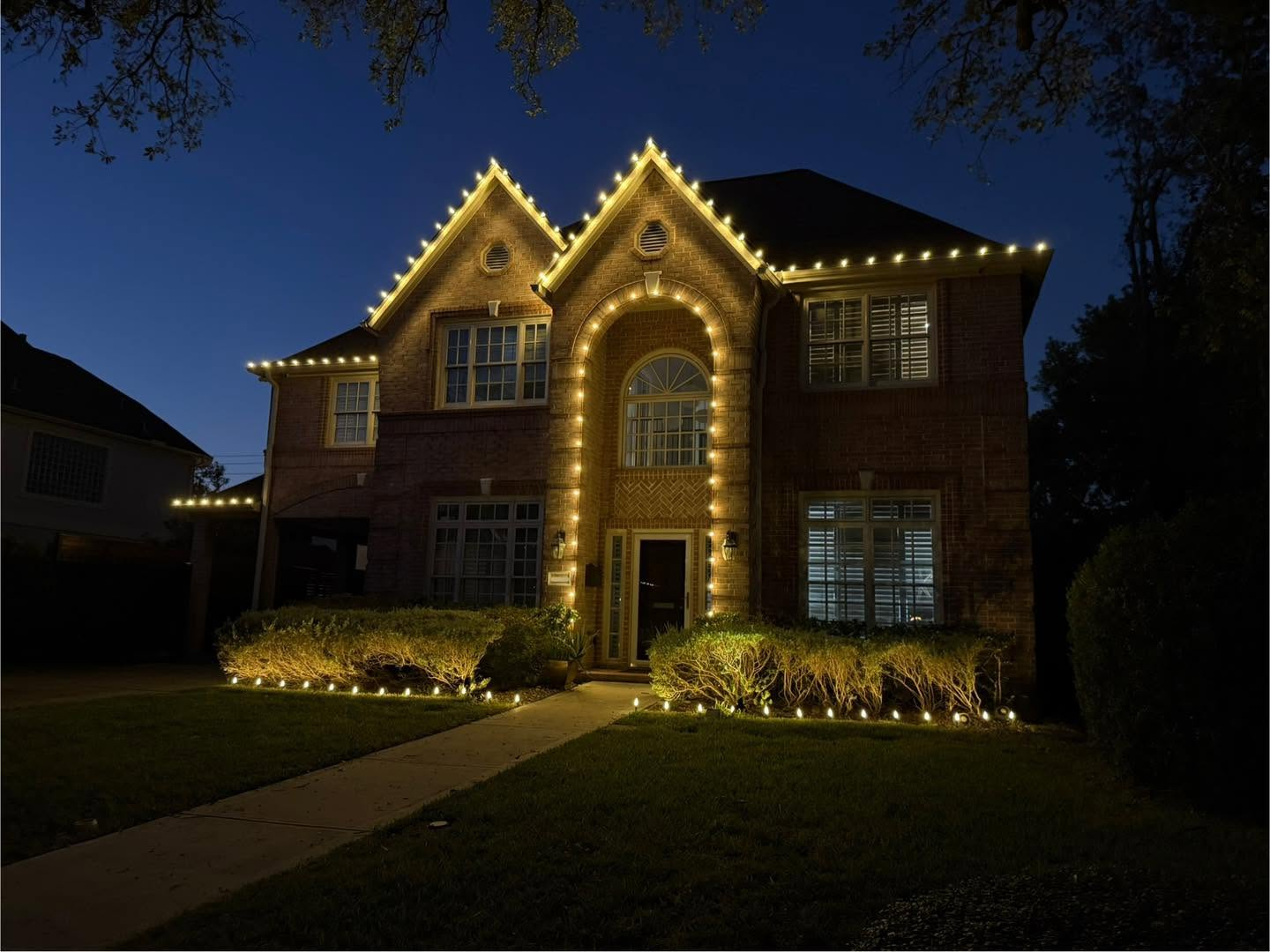 Brick two-story Houston home with warm roofline lights and lit landscaping